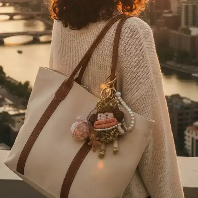 A person in a cream sweater carries a beige tote bag with brown straps, featuring a crochet doll keychain with pink, gold, and pearl details, against a sunset river backdrop.