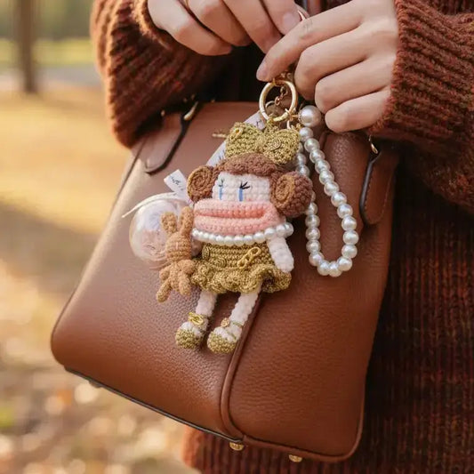 Hands holding a brown leather bag, adorned with a delicate crochet doll keychain featuring pink and gold clothing, paired with a pearl chain decoration, set against an autumn outdoor background.
