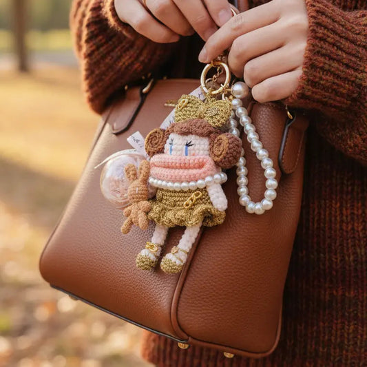 Hands holding a brown leather bag, adorned with a delicate crochet doll keychain featuring pink and gold clothing, paired with a pearl chain decoration, set against an autumn outdoor background.